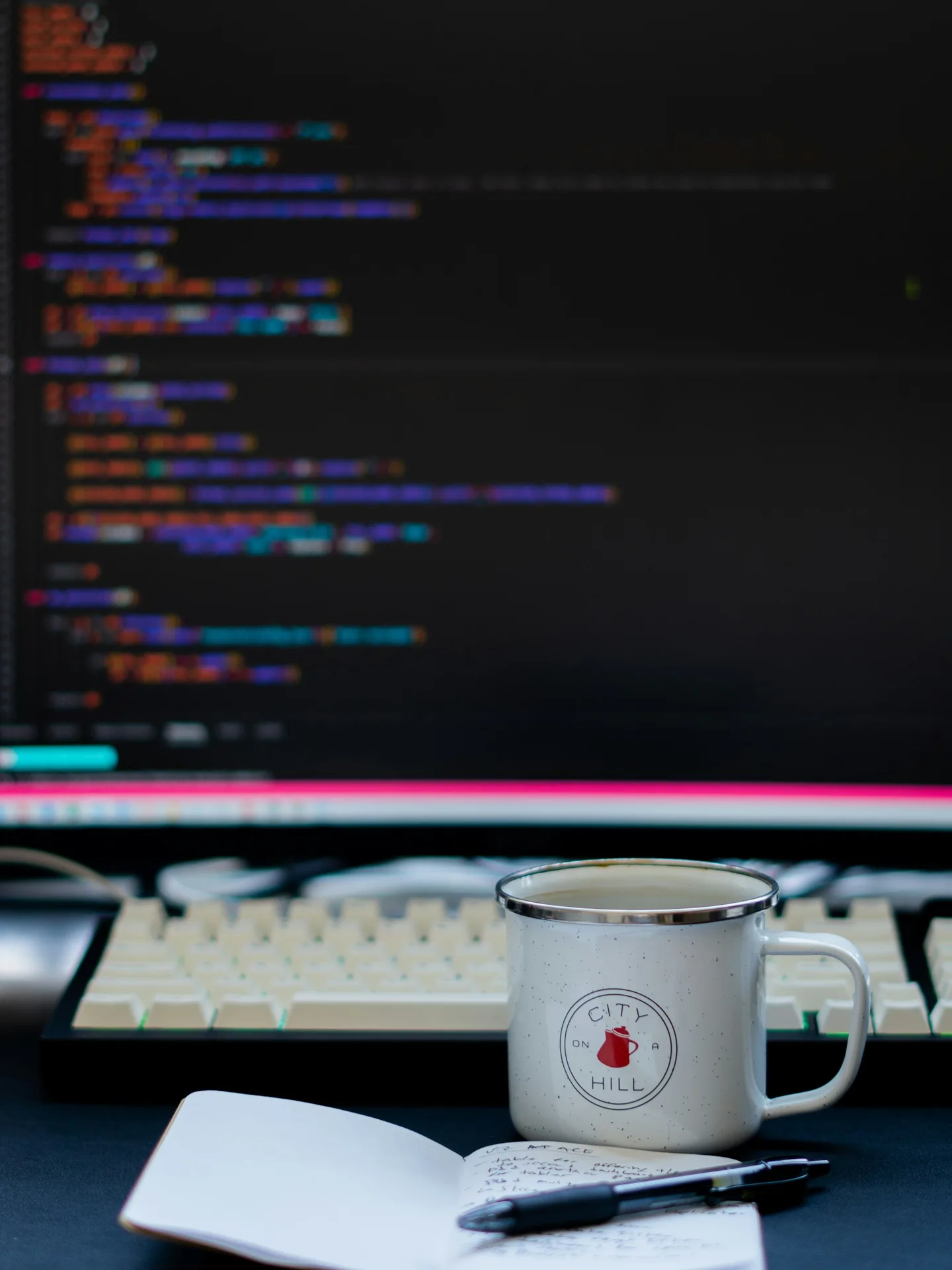Monitor con código de colores, teclado y una taza de peltre junto a una libreta abierta
