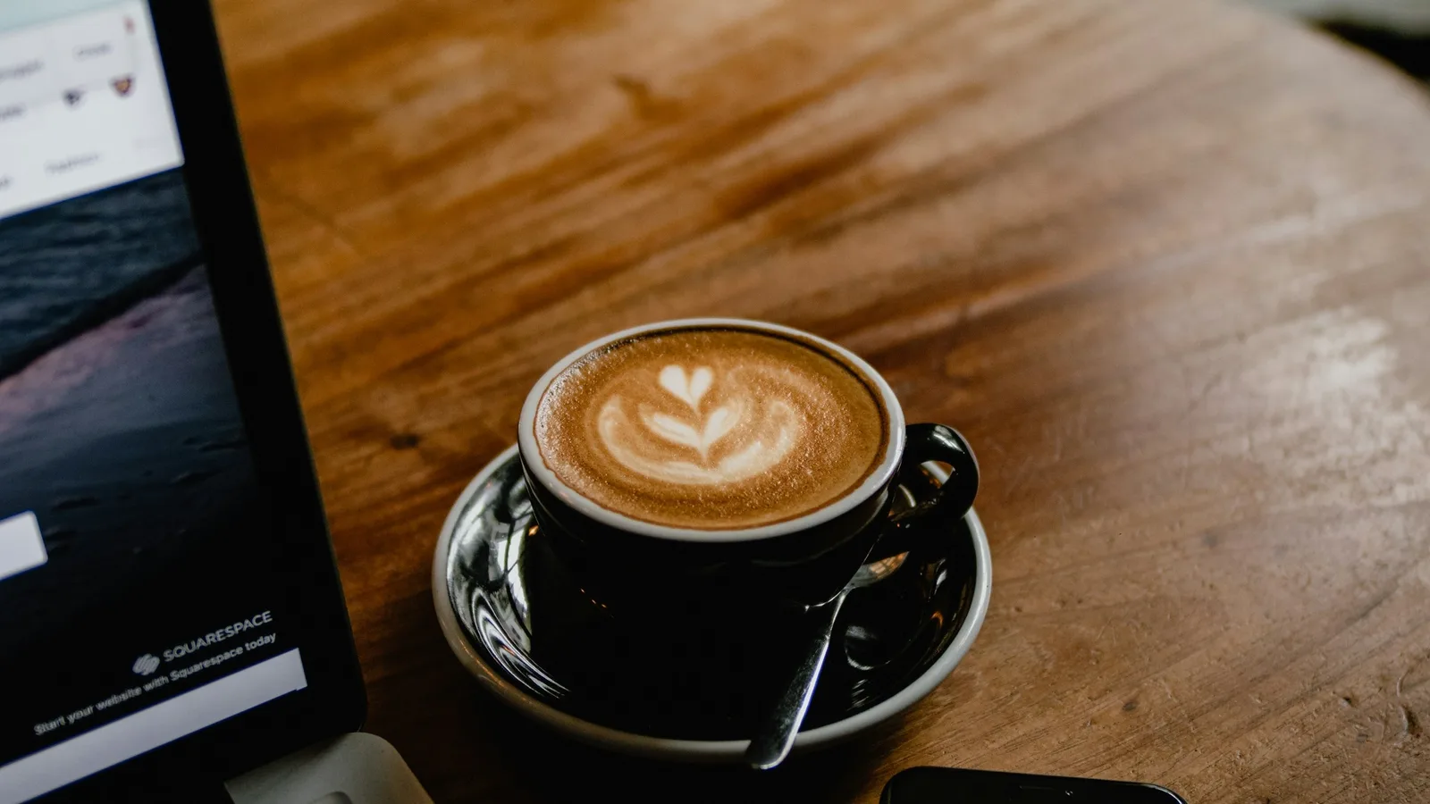 Taza de café con arte en la leche sobre una mesa de madera, junto a una laptop abierta