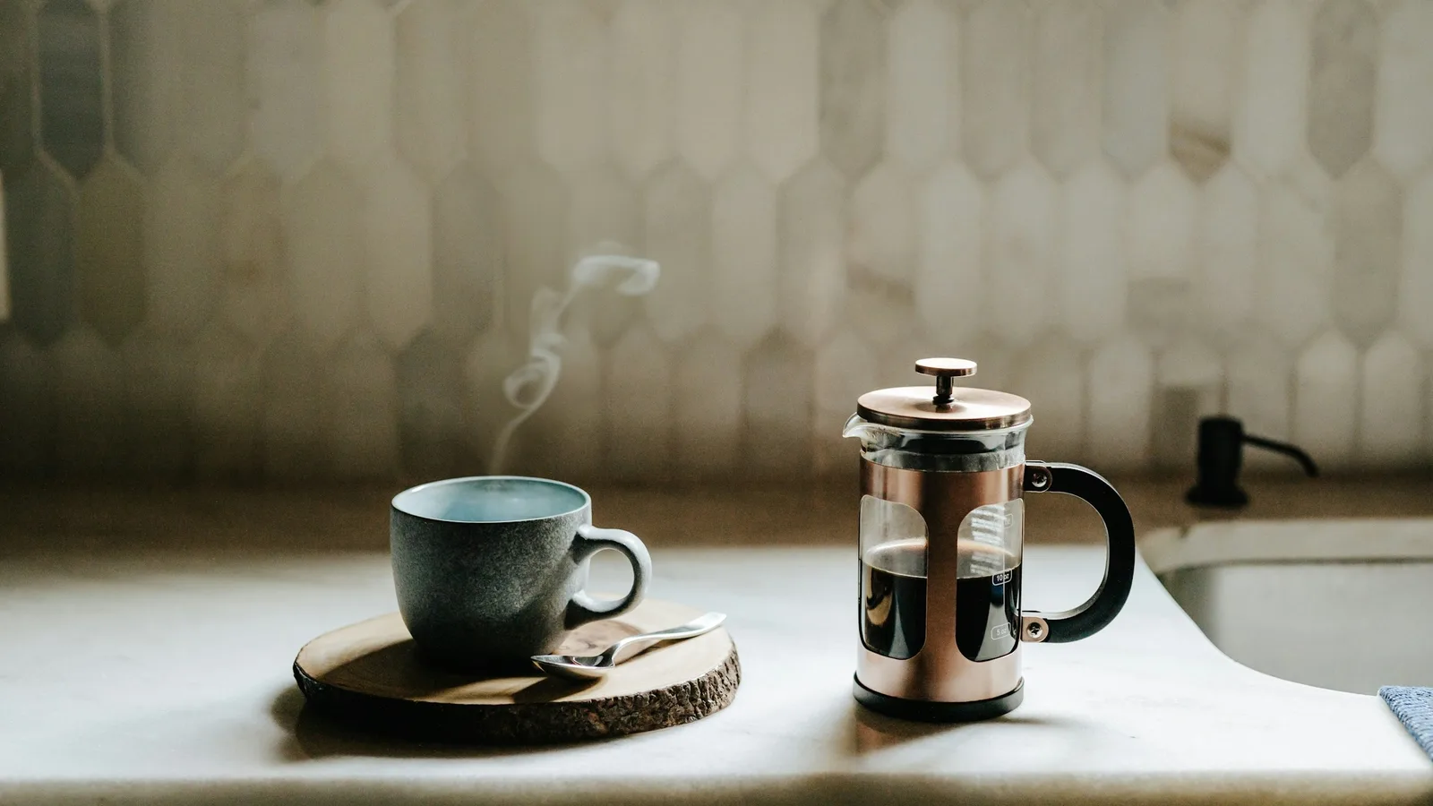 Prensa francesa de vidrio y metal con café, junto a una taza humeante en una barra de cocina